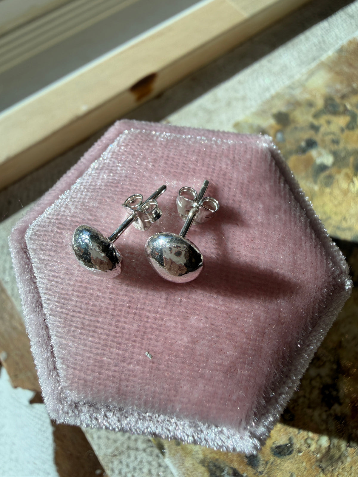 A pair of silver button stud earrings with round polished fronts and butterfly backs, displayed on a pink velvet hexagonal jewelry box.