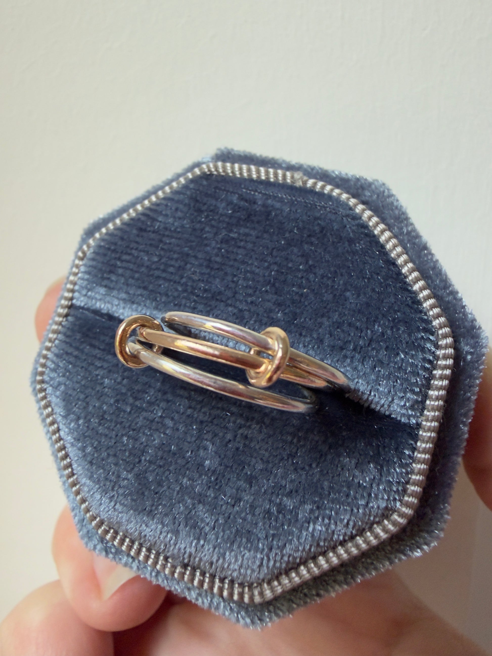 A close-up of a ring made from 2 silver ring and 9ct yellow gold ring interlocked by 2 small gold loops displayed inside an octagonal blue velvet ring box held by a hand.