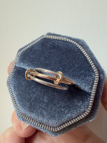 A close-up of a ring made from 2 silver ring and 9ct yellow gold ring interlocked by 2 small gold loops displayed inside an octagonal blue velvet ring box held by a hand.