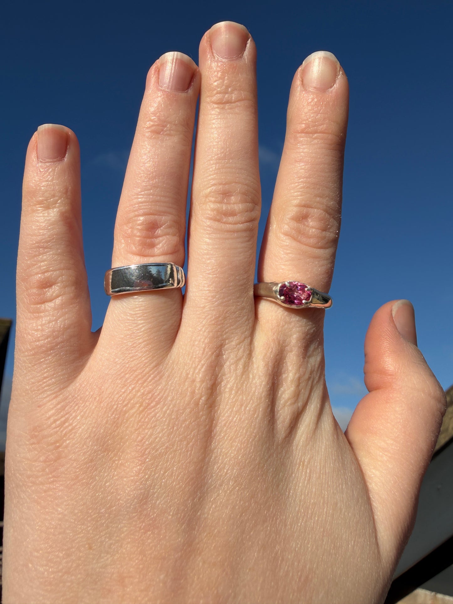 Hand wearing two rings, slim pink topaz signet and plain banner ring, with gemstones against a blue background
