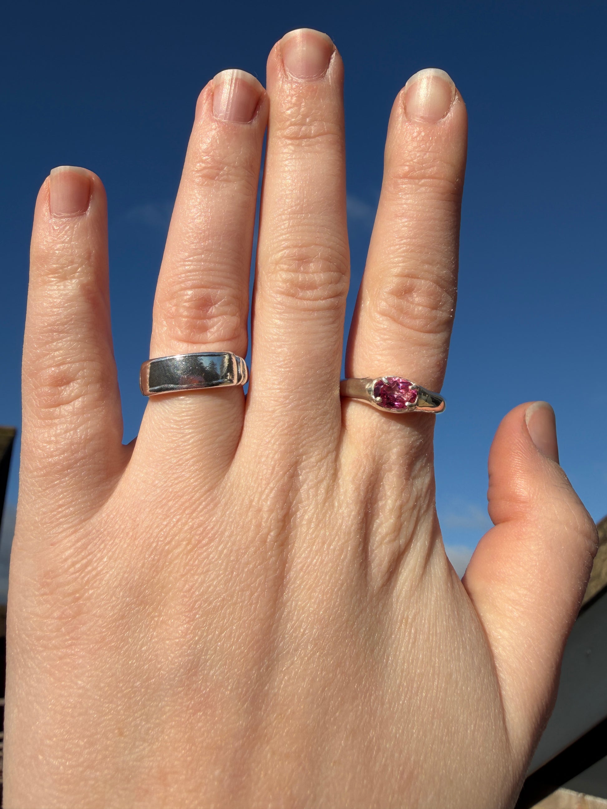 Hand wearing two rings, slim pink topaz signet and plain banner ring, with gemstones against a blue background