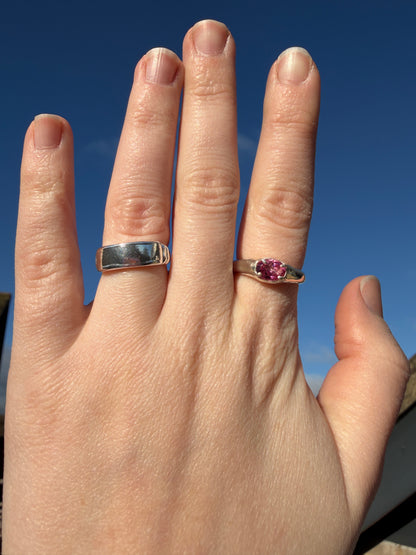 Hand wearing two rings, slim pink topaz signet and plain banner ring, with gemstones against a blue background