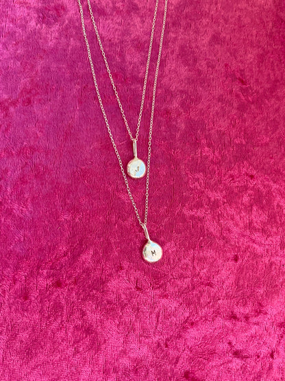 Two silver necklaces with round silver letter pendants on a pink textured fabric background