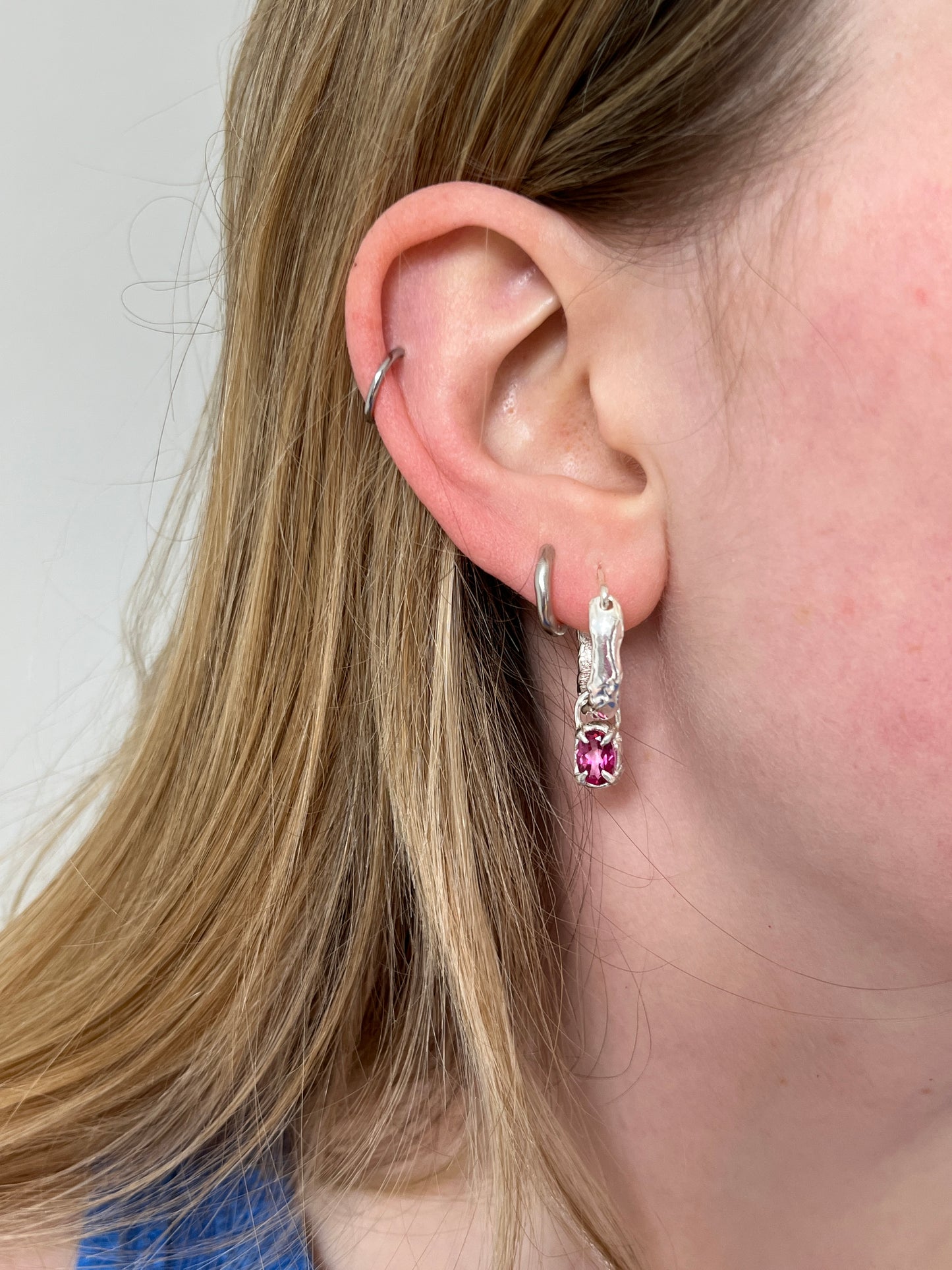 Close-up of an ear wearing a silver earring with a pink gemstone on a neutral background.
