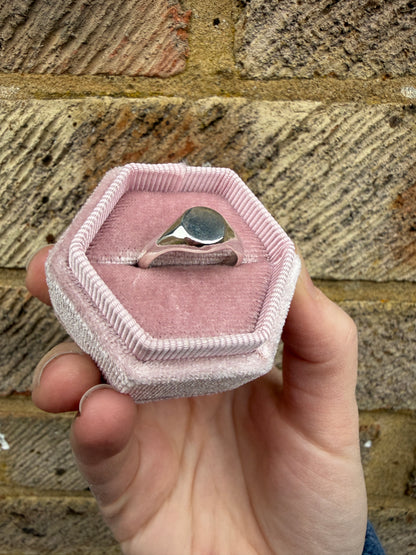 The silver mini signet pictured in a pink ring box with a brick background