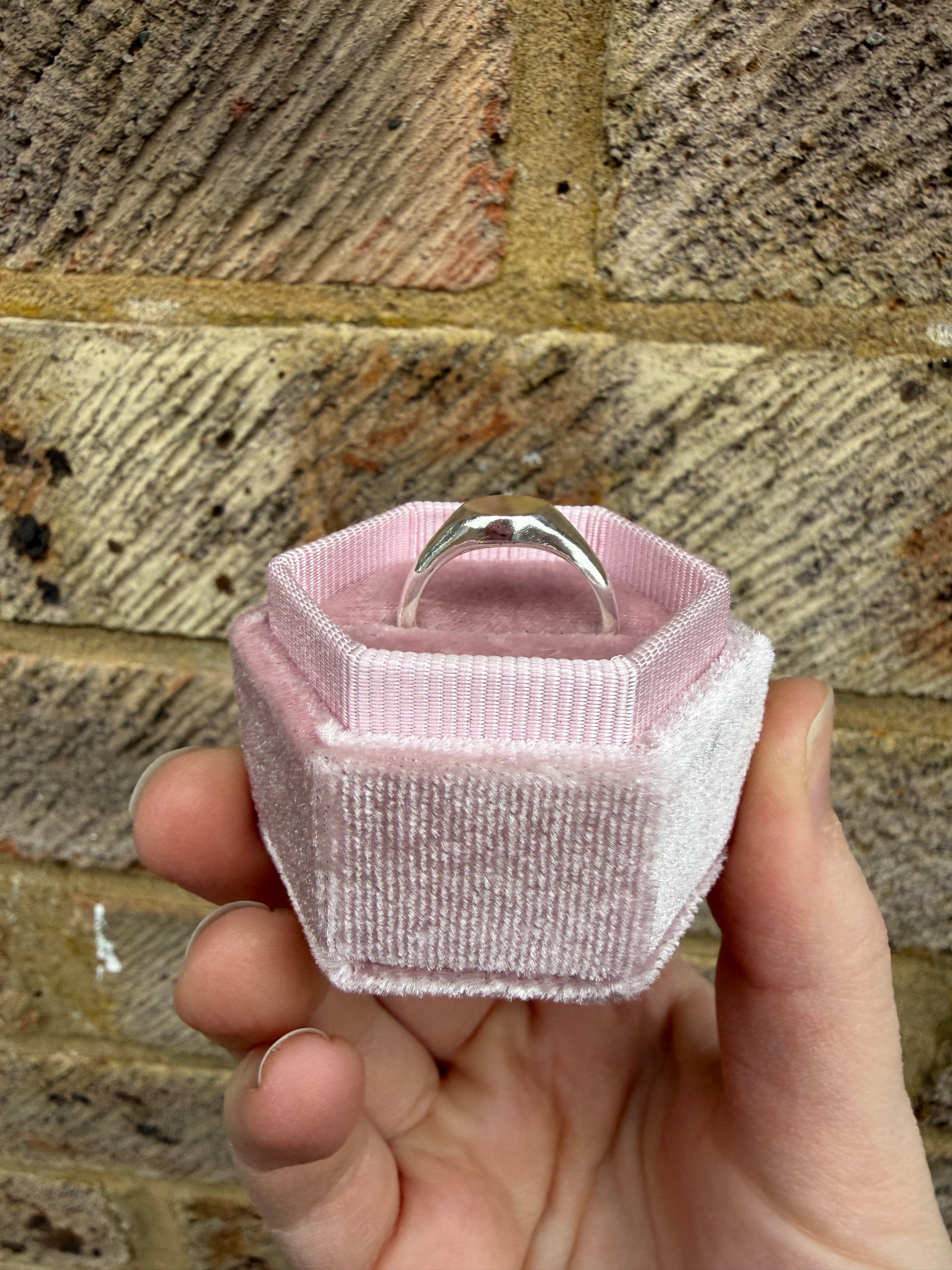 The silver mini signet pictured from the side in a pink ring box with a brick background