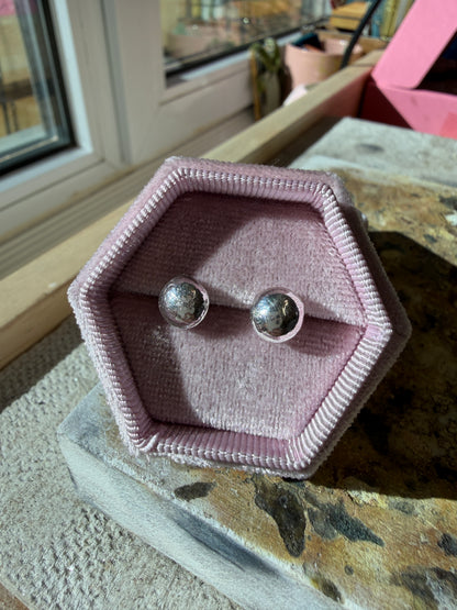 Molten sterling silver button stud earrings, pictured in a pink velvet jewellery box sat on a workbench.