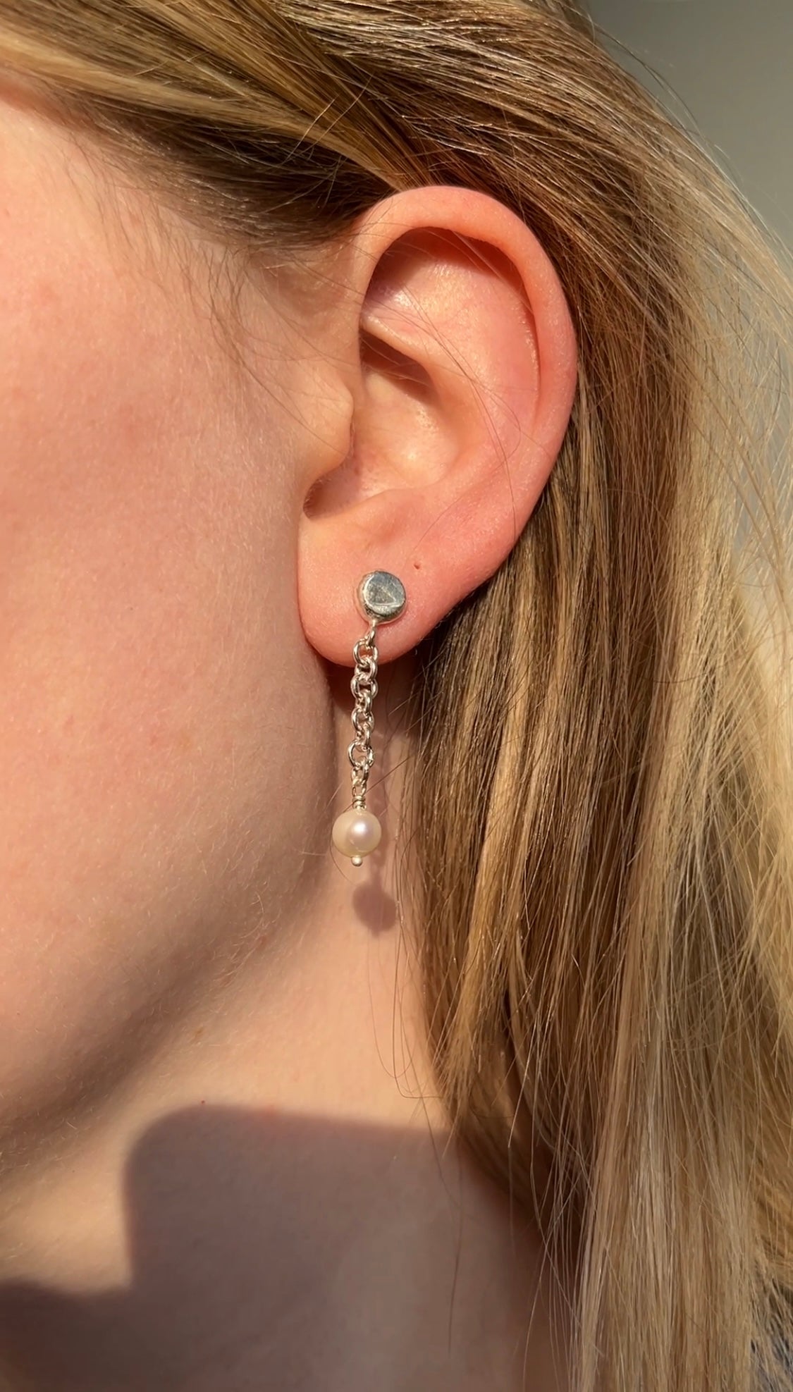 A close-up image of a person's ear wearing a silver stud pearl chain drop earring.
