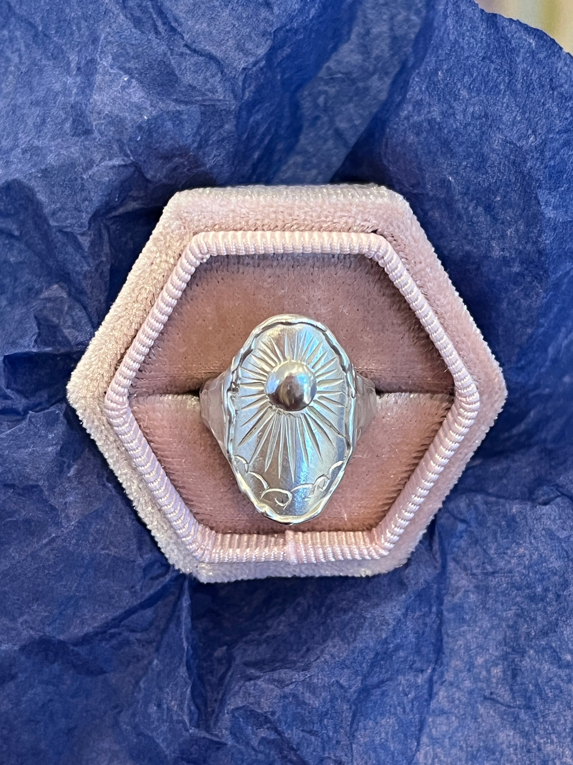 A sterling silver ring with an intricate beaming sun engraving and molten band design, displayed in a pink hexagonal box with a blue background.