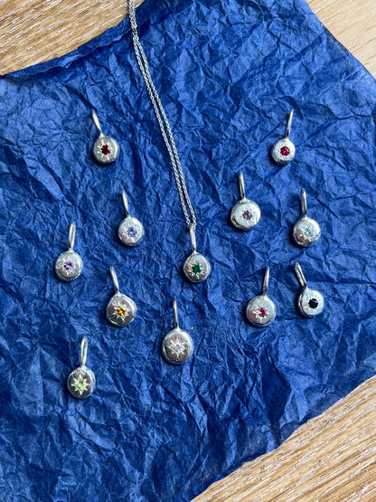 silver pendants with each months birthstone pictured on a blue paper background.